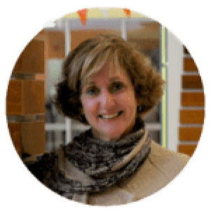 A woman with short curly hair is smiling, wearing a scarf and standing indoors in front of brick columns.