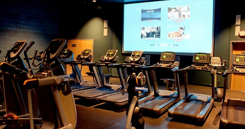 A row of treadmills facing a large screen in a dimly lit gym.
