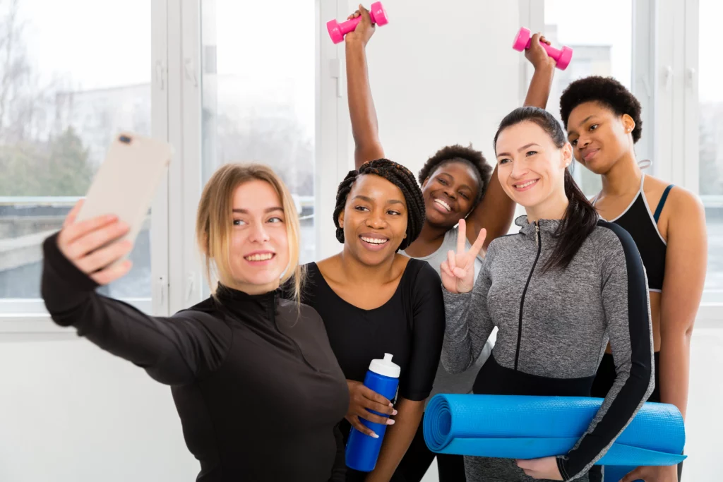 Five women in workout attire take a group selfie in a bright room, with yoga mats and dumbbells, smiling and posing.